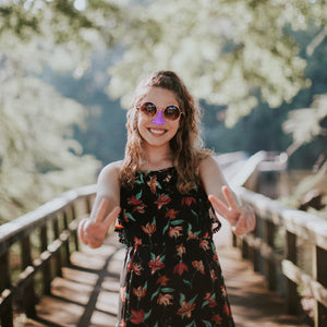 Young female in an outdoor scene wearing purple Nöz sunscreen stick on nose while giving two peace signs