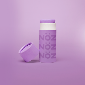 Purple Nöz sunscreen stick with cap off, tilted against base of stick. All against a pale purple gradient background. 