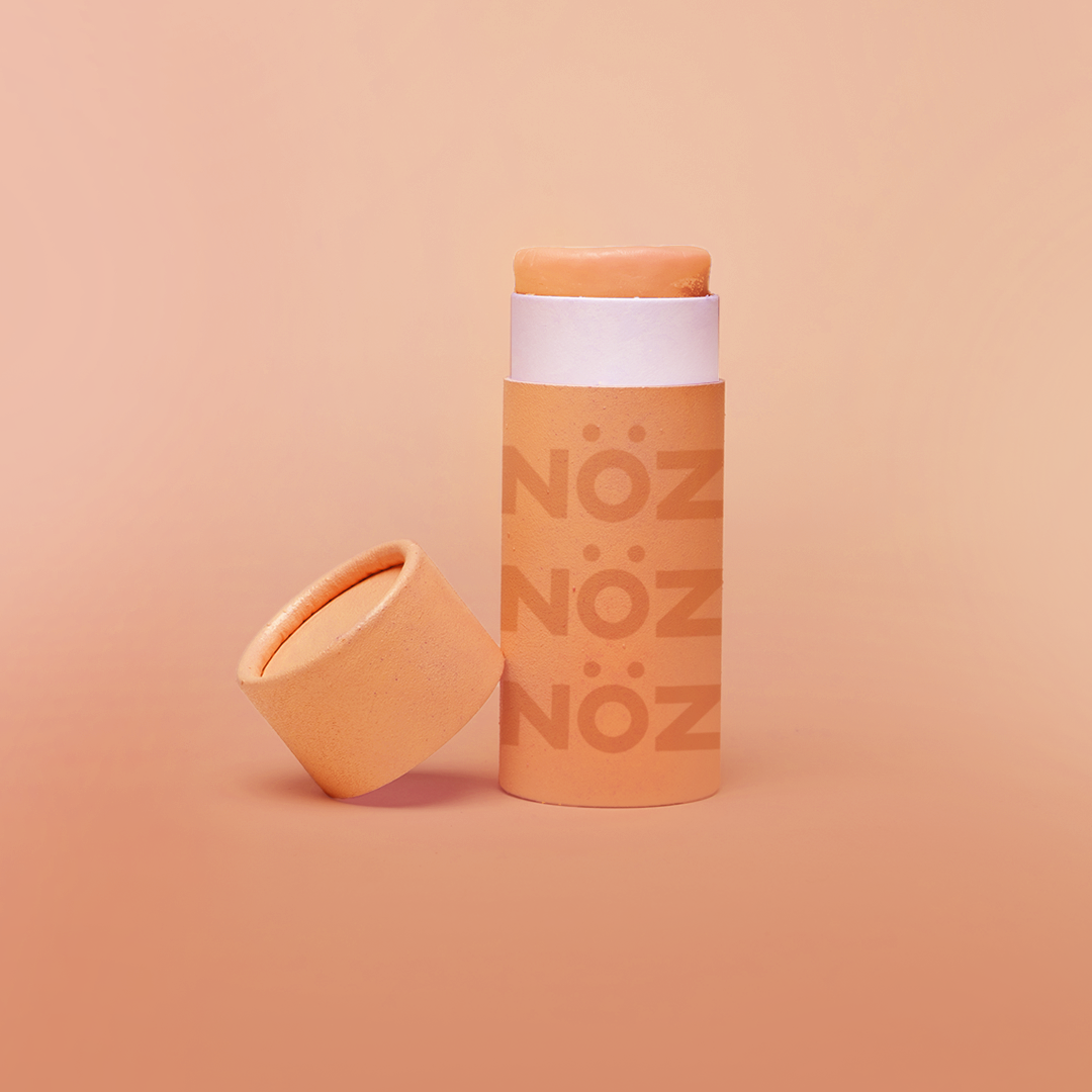 Orange Nöz sunscreen stick with cap off, tilted against base of stick. All against a pale orange gradient background. 