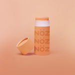 Load image into Gallery viewer, Orange Nöz sunscreen stick with cap off, tilted against base of stick. All against a pale orange gradient background. 
