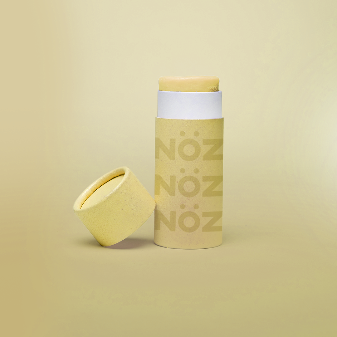 Yellow Nöz sunscreen stick with cap off, tilted against base of stick. All against a pale yellow gradient background. 