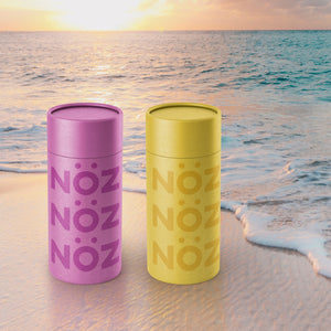 Purple and yellow Nöz sunscreen sticks against an ocean and sand background at sunset. 